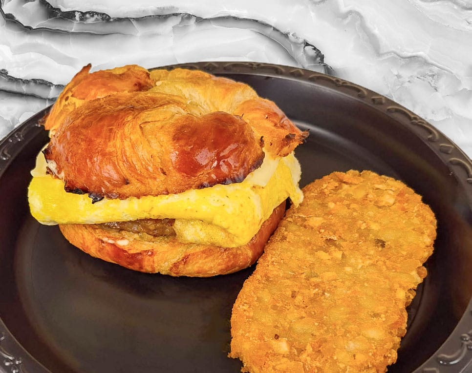 Croissant breakfast sandwich with egg and cheese on a dark plate, plus a crispy fried nugget beside it.
